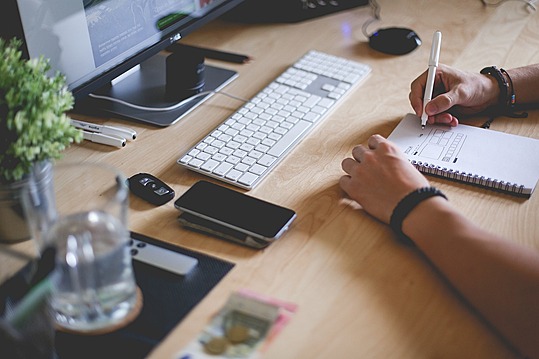 Home office desk setup with man writing on a notepad.